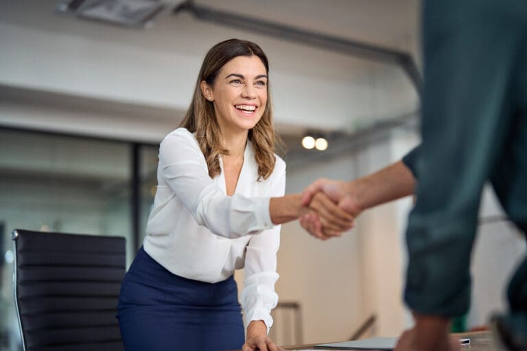 Happy mid aged business woman manager handshaking greeting client in office. Smiling female executive making successful deal with partner shaking hand at work standing at meeting table.